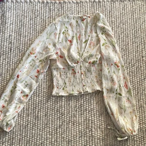Blommig blus med knytband - Säljer en superfin blommig blus med långa ärmar och knytband vid ärmsluten. Blusen har en v-ringad design och smockdetaljer i midjan som ger en snygg passform. Perfekt för vår och sommar!
