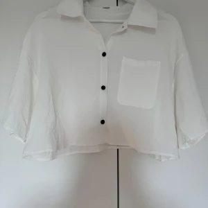 Vit croppad skjorta med knappar - Säljer en superfin vit croppad skjorta med svarta knappar. Den har en bröstficka och är perfekt för sommardagar. Skjortan har en lös passform och korta ärmar, vilket gör den både bekväm och stilren. Perfekt för en avslappnad look!