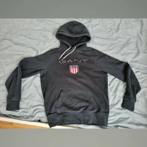 Mörkgrå hoodie från GANT - Säljer en snygg och bekväm hoodie från GANT. Den har en klassisk design med en stor ficka fram och justerbar huva med vita snören. Perfekt för en avslappnad stil eller kyliga dagar. GANT-loggan och ett stilrent emblem pryder framsidan.