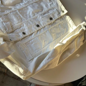 Vit jeansjacka från The Blue Uniform - Säljer en snygg vit jeansjacka från The Blue Uniform. Jackan har en klassisk design med knappar framtill och två bröstfickor. Perfekt för vår och sommar, och den har långa ärmar med knappar vid ärmsluten. Den är i bra skick och redo för nya äventyr!