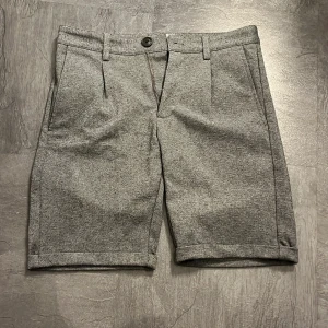 Grå shorts från Lindbergh - Säljer ett par stilrena grå shorts från Lindbergh. De har en klassisk design med dragkedja och knapp framtill. Shortsen är perfekta för både vardag och lite mer uppklädda tillfällen. De har en snygg passform och är gjorda i ett bekvämt material.