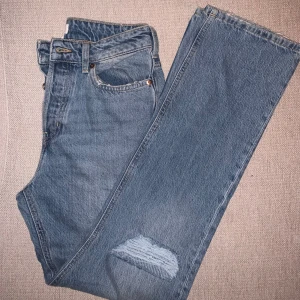 Blå jeans med slitningar - Säljer ett par snygga blå jeans med slitningar på ena benet. De har en klassisk rak design med knappar istället för dragkedja. Aldrig använda 