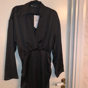 Svart klänning från Zara - Säljer en elegant svart klänning från Zara. Den har en stilren design med långa ärmar och en omlott-look framtill. Materialet känns lyxigt och har en fin glans. Storlek M, helt ny 