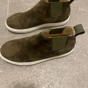 Bruna mocka boots - Säljer ett par snygga gröna boots i mocka. De har en vit sula och elastiska paneler på sidorna för enkel på- och avtagning. Perfekta för höst och vår, och de ger en stilren look till vilken outfit som helst.