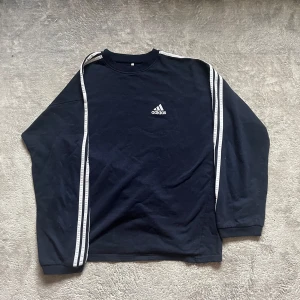 Svart långärmad tröja från Adidas - Säljer en svart långärmad tröja från Adidas med vita ränder längs ärmarna och det klassiska Adidas-loggan på bröstet. Perfekt för en sportig look eller bara för att chilla i. Tröjan är i bra skick och superbekväm!