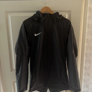 Svart vindjacka från Nike - Säljer en svart vindjacka från Nike i storlek s Jackan har en dragkedja framtill och en praktisk huva. Perfekt för blåsiga dagar och har en stilren design med Nike-loggan på bröstet. Passar bra till både träning och vardagsbruk. Aldrig använd.