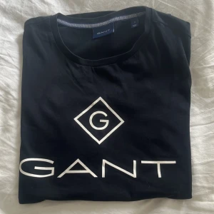 Svart t-shirt från GANT - Säljer en snygg svart t-shirt från GANT med deras ikoniska logga tryckt i vitt på framsidan. Perfekt för en casual look och passar till det mesta. T-shirten är i bra skick och redo för en ny ägare!