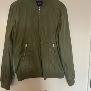 Olivgrön bomberjacka från GANT - Säljer en snygg olivgrön bomberjacka från GANT i storlek M. Jackan har en stilren design med dragkedja framtill och två praktiska fickor med dragkedjor. Perfekt för höst och vår!