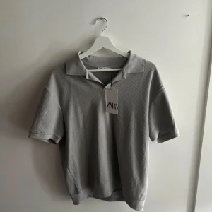 Grå pikétröja från Zara - Säljer en stilren grå pikétröja från Zara. Den har en klassisk krage och korta ärmar, perfekt för en avslappnad men ändå snygg stil. Materialet är ribbat. Helt ny med prislapp kvar. Originalpris 300kr
