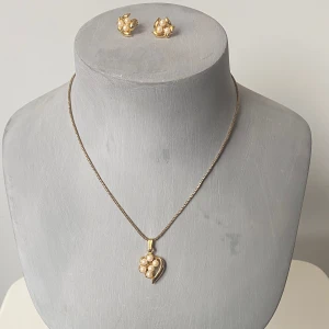 Guldhalsband och örhängen med pärlor - Säljer ett elegant set med ett guldhalsband och matchande örhängen. Halsbandet har en fin kedja med ett hängsmycke format som ett hjärta, dekorerat med små pärlor. Örhängena är i samma stil med guldblad och pärlor i mitten. Okänd metall 