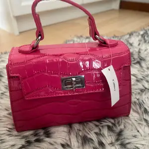 Säljer en superfin rosa oanvänd handväska med krokodilmönster. Den har ett snyggt metallspänne framtill och ett praktiskt handtag. Perfekt för att liva upp vilken outfit som helst!  Ny pris: 299kr