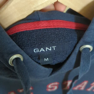 Mörkblå hoodie från GANT - Säljer en snygg mörkblå hoodie från GANT med röd text och ett coolt universitetsmärke på bröstet. Den har en klassisk känguruficka och justerbar huva med snören. Använd några gånger men den är så gott som ny och väl omhändertagen.