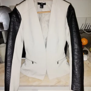 Vit och svart jacka från H&M - Säljer en snygg vit och svart jacka från H&M. Den har en cool design med vita paneler och svarta skinnärmar. Jackan har dragkedjor framtill och en stilren look. Perfekt för både vardag och fest!