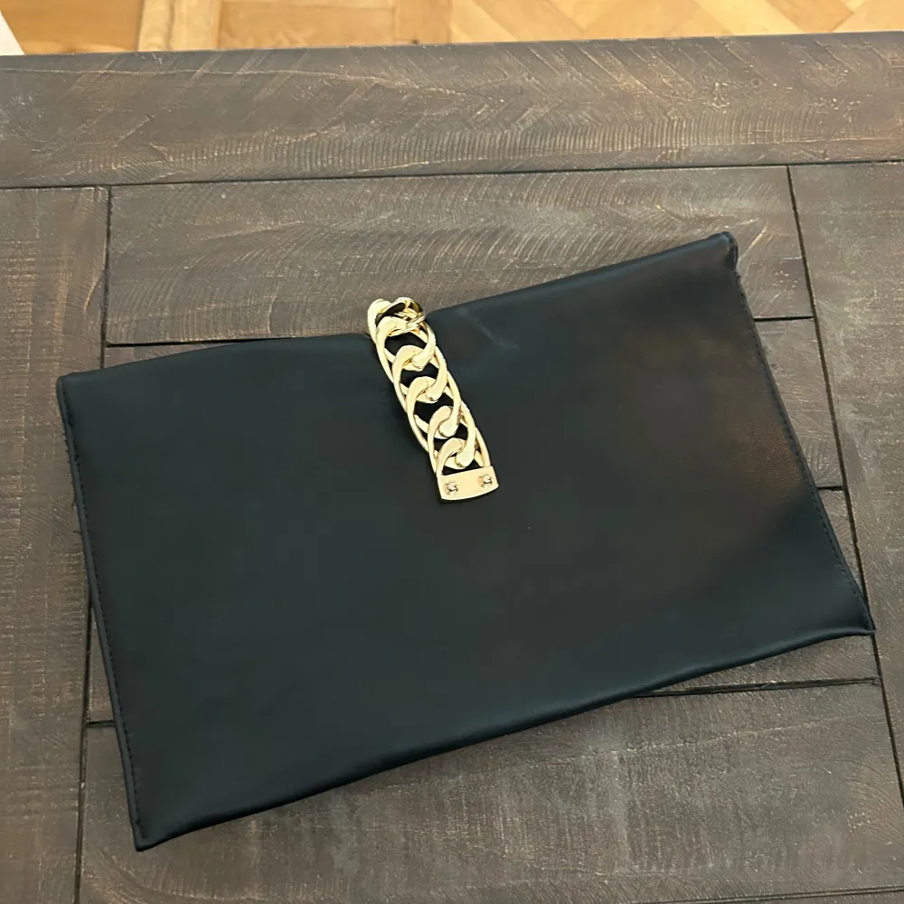 Snygg svart kuvertväska i syntetmaterial med en elegant guldkedja som detalj. Väskan har en praktisk dragkedja inuti för extra säkerhet. Perfekt för festliga tillfällen eller en utekväll när du vill ha något stilrent och enkelt. . Laukut & Käsilaukut.
