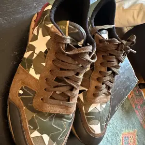 Säljer ett par snygga Valentino Garavani sneakers i kamouflagemönster. Skorna har en cool mix av gröna och bruna nyanser med vita stjärnor. De är gjorda i en kombination av skinn och mocka, och har de ikoniska rockstud-detaljerna på hälen. Perfekta för att ge din outfit en edgy touch!