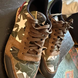 Valentino Garavani sneakers med kamouflagemönster - Säljer ett par snygga Valentino Garavani sneakers i kamouflagemönster. Skorna har en cool mix av gröna och bruna nyanser med vita stjärnor. De är gjorda i en kombination av skinn och mocka, och har de ikoniska rockstud-detaljerna på hälen. Perfekta för att ge din outfit en edgy touch!