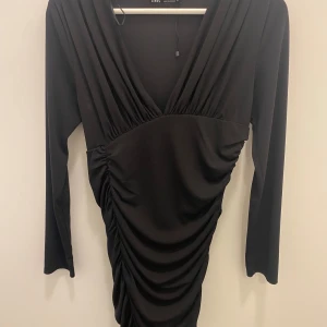 Svart klänning från Zara - Säljer en elegant svart klänning från Zara. Den har en v-ringad design med långa ärmar och en snygg rynkad detalj baktill och på sidorna som ger en smickrande passform. Perfekt för en festlig kväll eller en utekväll med vänner! Klänningen är i jättebra skick endast använd en gång!