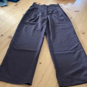 Bruna byxor från Monki - Säljer ett par snygga bruna byxor från Monki. De är högmidjade och har en vid passform som ger en skön och avslappnad stil. Byxorna är gjorda av hållbart material och har knappar i midjan. Perfekta för både vardag och fest!