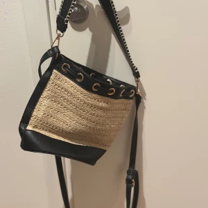 Beige och svart axelväska med kedjedetalj - Axelväska i beige och svart med flätat mönster och kedjedetalj på axelremmen. Väskan har en praktisk dragskoöppning med metallöljetter och en liten ficka inuti. Köpt för flera år sedan på en marknad dock aldrig använt den. 