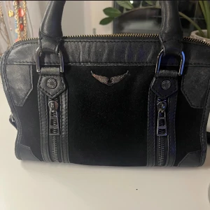 Svart axelväska med dragkedja - Snygg svart axelväska i skinn med coola detaljer. Väskan har en praktisk dragkedja upptill och två dekorativa dragkedjor framtill. Den har en avtagbar axelrem och robusta handtag. Perfekt för att ge din outfit en edgy touch. Jag har även en dustbag till den och tag.