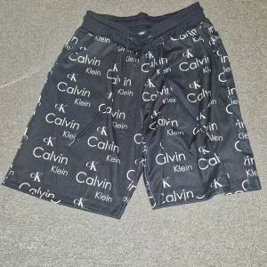 Svarta shorts från Calvin Klein - Snygga svarta shorts från Calvin Klein med ett stilrent mönster av märkets logotyp över hela tyget. De har en elastisk midja med snörning för bästa passform. Perfekta för en avslappnad stil.