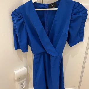 Blå klänning med volangdetaljer - Snygg blå klänning från A/X med volangdetaljer på ärmarna och en djup v-ringning. Perfekt för att sticka ut med sin unika design. Klänningen har en dragkedja i ryggen för enkel påklädning.