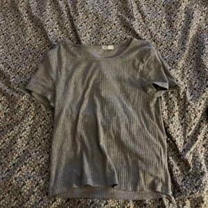 Grå ribbad t-shirt från C - Säljer en stilren grå ribbad t-shirt från märket C. Den har en klassisk rund hals och korta ärmar, perfekt för en avslappnad look. Passar bra till jeans eller kjol för en enkel och snygg outfit.