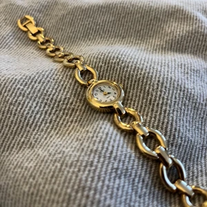 Liten vintage klocka - Liten vintage klocka i guldfärg. Metallen på själva uret är i rostfritt stål, även metallen på klockan är det. Vet inte om klockan är guldpläterad, men den har inte tappat någon guldfärg trots att modellen är mycket gammal. Batteriet i uret har tyvärr slutat funka, men går säkert att byta ut om man vill att den ska funka igen! Omkretsen är 19 cm, men det går att knäppa bort en länk och då är omkretsen 17,5 cm. Aldrig använd då plasten på baksidan av uret fortfarande är kvar!