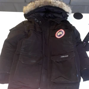Svart parkajacka från Canada Goose - Säljer en svart parkajacka från Canada Goose med pälsfodrad huva. Jackan har en broderad logotyp på bröstet och flera praktiska fickor. Perfekt för kalla vinterdagar. 🖤