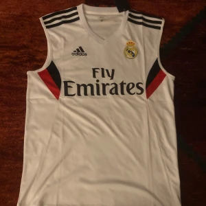 Vit ärmlös fotbollströja. finns i alla storlekar  - Säljer en vit ärmlös fotbollströja med Real Madrid-logga och Fly Emirates-tryck. Tröjan har svarta och röda detaljer vid sidorna och tre svarta ränder på axlarna. Perfekt för fotbollsfans!