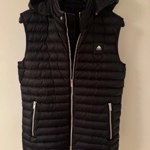 Svart dunväst från Moose Knuckles - Säljer en stilren svart dunväst från Moose Knuckles. Västen har en praktisk dragkedja framtill och två fickor med dragkedjor. Perfekt för kyligare dagar med sin quiltade design och avtagbar huva. Nypris 6000kr säljer för 4500. Notera att storleken är S men att den passar större. Be om mått eller bild på passform. Tags finns 