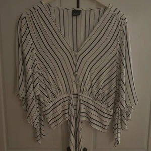 Randig blus från Gina Tricot - Säljer en stilren vit blus med svarta ränder från Gina Tricot. Blusen har en v-ringad design med knappar framtill och vida ärmar. Nästan helt oanvänd🥰