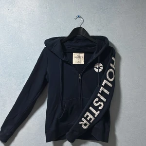 Mörkblå zip-hoodie från Hollister - Säljer en riktigt snygg zip up-hoodie i mörkblå/marinblå färg från hollister. Ärmarna är detaljerade i Hollister text vilket ger en snygg konstrast till tröjan. Tröjan är riktigt snygg men behöver tyvärr sälja den pga att den är lite liten. Den är i storlek S och den enda defekten jag kunde hitta var vid vänstra fickan, en liten fläck men har inte testat att tvätta bort den och den syns endast i riktigt strakt ljus. Storlek S från Hollister, skick 8,5/10 (en liten fläck som är svår synlig) 