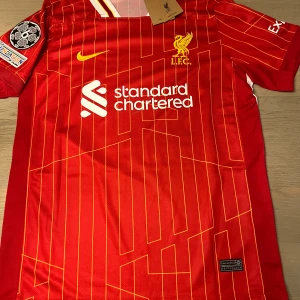 Röd Liverpool fotbollströja - Säljer en röd Liverpool fotbollströja från Nike med gula detaljer och klubbens emblem på bröstet. Tröjan har korta ärmar och ett randigt mönster. Den har även tryck med 'M. Salah' och nummer 11 på ryggen. Perfekt för fans av laget!