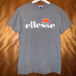 Grå t-shirt från Ellesse - Snygg grå t-shirt från Ellesse med deras ikoniska logga i vitt och orange på bröstet. Perfekt för en avslappnad stil. T-shirten har en klassisk passform och är kortärmad.