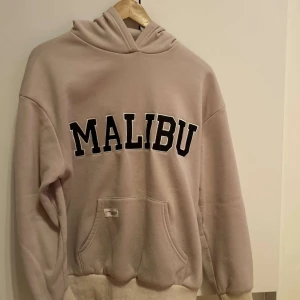 Beige hoodie med texten MALIBU - En mysig hoodie med tryck från shein. Fint skick förutom ett litet brännmärke på baksidan som går att fixa ihop. Den är ganska oversized 