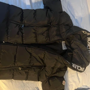 Svart dunjacka från Moncler - Säljer en snygg svart dunjacka från Moncler med dragkedja och logotypdetaljer på kragen. Jackan har en quiltad design och är perfekt för kyligare väder. Den har långa ärmar och praktiska fickor med dragkedjor.