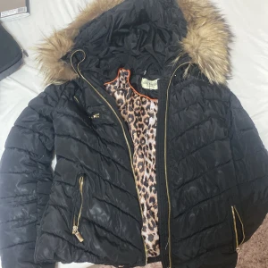 Svart dunjacka från Roco Baroco - Snygg svart dunjacka från Roco Baroco med leopardmönstrat foder och pälskantad huva. Jackan har dragkedja och praktiska fickor med dragkedjor. Perfekt för kyliga dagar.