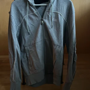 Grå huvtröja från Boss - Säljer en stilren grå huvtröja från Boss med dragkedja och justerbar huva. Tröjan har långa ärmar och praktiska fickor framtill. Perfekt för en avslappnad look.