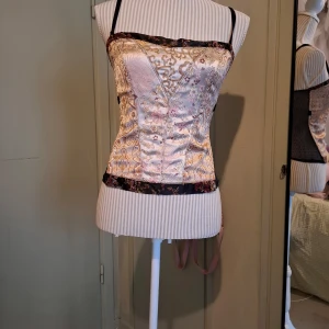 Mönstrad korsett från H&M - Säljer en elegant korsett från H&M med ett vackert blommigt mönster i guld, rosa och lila nyanser. Korsetten har justerbara axelband och en dragkedja i sidan för enkel påklädning. Perfekt för att ge en stilfull touch till din outfit.