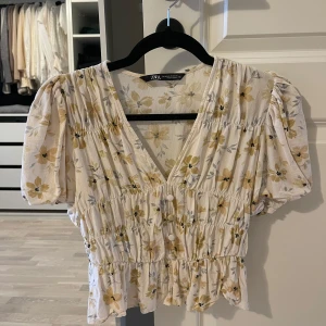 Blommig blus från Zara - Säljer en söt blommig blus från Zara i ljus beige med gula och grå blommor. Blusen har korta puffärmar och en v-ringad design med knappar framtill. Perfekt för vår och sommar!