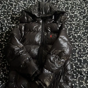 Ralph Lauren jacka - Snygg svart dunjacka från Polo Ralph Lauren med glansig finish. Jackan har en dragkedja framtill och en huva för extra värme. Perfekt för kyliga dagar. Använd max 2-3 gånger. Säljes för jag använder andra.