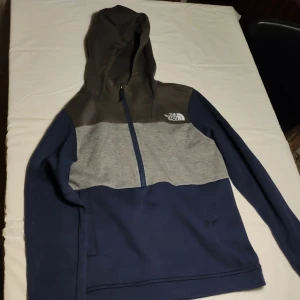 Grå och blå hoodie från The North Face - Säljer en snygg grå och blå hoodie från The North Face. Tröjan har en dragkedja framtill och en bekväm huva. Perfekt för kyliga dagar och avslappnade stunder. Logotypen är tryckt på bröstet.