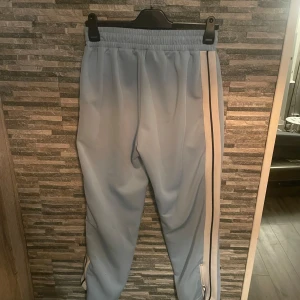 Ljusa byxor från Palm Angels - Snygga ljusblå byxor från Palm Angels med vita och svarta ränder längs sidorna. Byxorna har en elastisk midja och är tillverkade i 100% polyester. Pris kan diskuteras