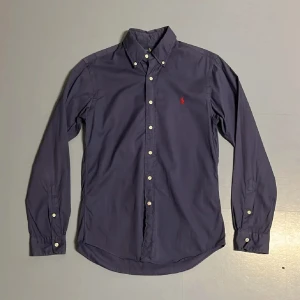 Mörkblå skjorta från Ralph Lauren - Snygg mörkblå skjorta från Ralph Lauren i slim fit. Skjortan har långa ärmar och knappar framtill. Den är tillverkad i ett lätt twillmaterial och har en röd broderad logga på bröstet.