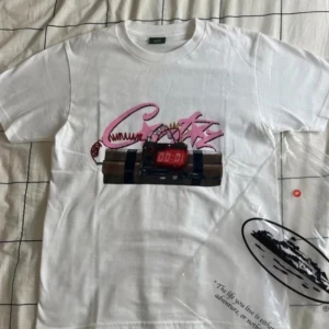 Corteiz T-Shirt - Säljer en vit cortiez t-shirt med ett unikt tryck av en digital klocka och graffiti-inspirerad text i rosa och svart. T-shirten har en klassisk rund hals och korta ärmar. Perfekt för en avslappnad stil.