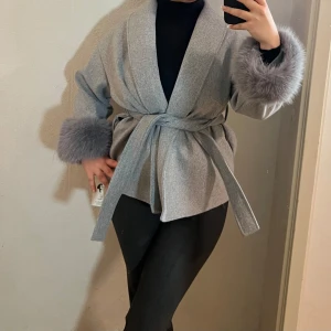 Grå kappa med fuskpälsdetaljer - Säljer en elegant grå kappa med fuskpälsdetaljer vid ärmsluten. Jackan har en omlott-design med knytband i midjan för en justerbar passform. Perfekt för kyligare dagar med sin stilrena look och mjuka material. Nypris 1800kr, har aldrig använt den:/ 