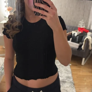 Svart topp från Monki - Säljer en svart topp från Monki med ett unikt texturerat mönster. Toppen har korta ärmar och en vågig kant vid ärmslut och nederkant. Perfekt för en stilren look. Tröjan är perfekt till vardags i kombination med ett par ljusa jeans! Tröjan säljs för att den inte används! 