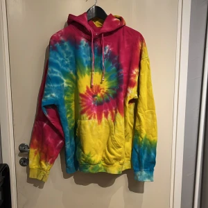 Färgglad tie-dye hoodie från Colortone - Säljer en färgglad tie-dye hoodie från Colortone i storlek L. Hoodien har en mix av rosa, blå, gula och lila färg i ett spiralmönster. Justerbar huva med snören. Perfekt för en avslappnad och färgstark stil!