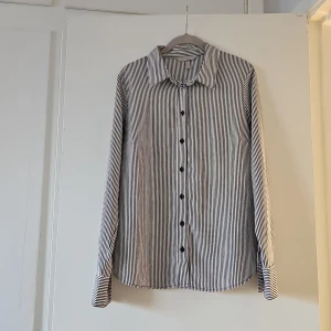 Svartvit randig skjorta från H&M - Snygg svartvit randig skjorta från H&M i storlek L. Skjortan har långa ärmar och knappar framtill. Tillverkad i 100% polyester vilket ger en lätt och bekväm känsla. Perfekt för en stilren look.